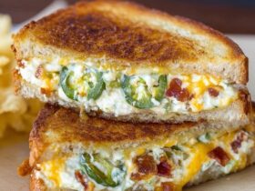 Jalapeño popper grilled cheese cut in half on a wooden board, golden-brown buttery toasted sourdough bread, cheesy melted cream cheese and cheddar stretchy pull, crispy bacon pieces, sliced pickled jalapeños visible in the gooey filling