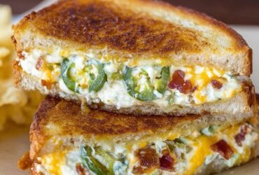 Jalapeño popper grilled cheese cut in half on a wooden board, golden-brown buttery toasted sourdough bread, cheesy melted cream cheese and cheddar stretchy pull, crispy bacon pieces, sliced pickled jalapeños visible in the gooey filling