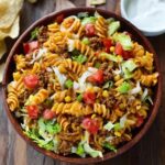 Taco pasta salad in a large white serving bowl, colorful rotini pasta mixed with seasoned ground beef, black beans, golden corn kernels, diced red tomato, shredded cheddar cheese, sliced green onions, crushed tortilla chips on top, creamy ranch-salsa dressing