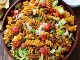 Taco pasta salad in a large white serving bowl, colorful rotini pasta mixed with seasoned ground beef, black beans, golden corn kernels, diced red tomato, shredded cheddar cheese, sliced green onions, crushed tortilla chips on top, creamy ranch-salsa dressing