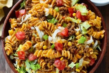 Taco pasta salad in a large white serving bowl, colorful rotini pasta mixed with seasoned ground beef, black beans, golden corn kernels, diced red tomato, shredded cheddar cheese, sliced green onions, crushed tortilla chips on top, creamy ranch-salsa dressing