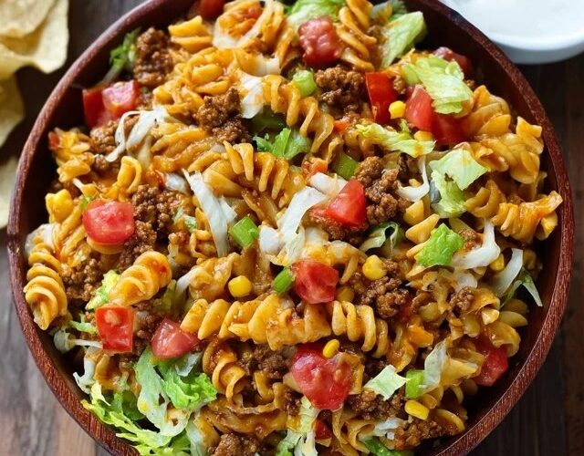 Taco pasta salad in a large white serving bowl, colorful rotini pasta mixed with seasoned ground beef, black beans, golden corn kernels, diced red tomato, shredded cheddar cheese, sliced green onions, crushed tortilla chips on top, creamy ranch-salsa dressing