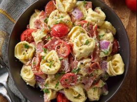 Tortellini salad with bacon in a large white serving bowl, plump cheese tortellini tossed with crispy bacon pieces, fresh baby spinach leaves, halved cherry tomatoes, sliced red onion, shredded parmesan, glossy Italian vinaigrette dressing coating everything, fresh basil leaves on top