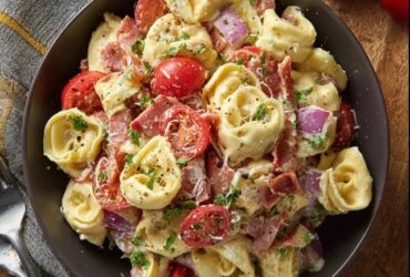 Tortellini salad with bacon in a large white serving bowl, plump cheese tortellini tossed with crispy bacon pieces, fresh baby spinach leaves, halved cherry tomatoes, sliced red onion, shredded parmesan, glossy Italian vinaigrette dressing coating everything, fresh basil leaves on top