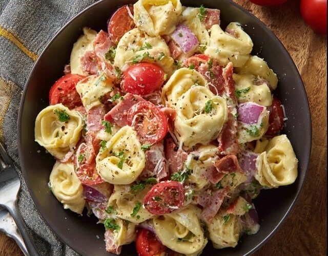 Tortellini salad with bacon in a large white serving bowl, plump cheese tortellini tossed with crispy bacon pieces, fresh baby spinach leaves, halved cherry tomatoes, sliced red onion, shredded parmesan, glossy Italian vinaigrette dressing coating everything, fresh basil leaves on top