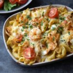 Shrimp pasta bake in a white ceramic baking dish, bubbly golden cheese-browned top revealing pink shrimp peeking through, penne pasta tossed in creamy tomato sauce, fresh basil leaves scattered, garlic bread on the side
