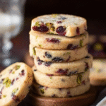 Cranberry pistachio shortbread cookies on a wire rack, pale buttery shortbread rounds with ruby-red dried cranberries and bright green chopped pistachios embedded throughout, festive holiday cookie tin in background