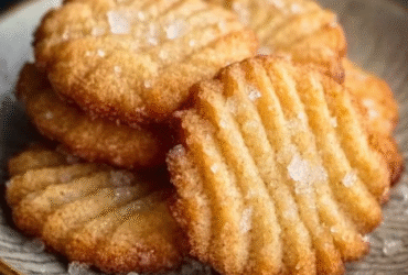 Danish butter cookies arranged in a vintage blue tin, golden-yellow piped swirl rosettes and pretzel shapes, pale buttery surface, sandy sugar crystals on top, cookie tin lid lifted to reveal contents