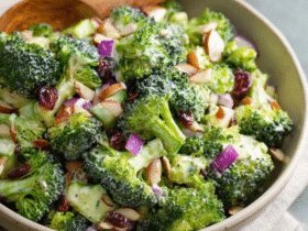 Broccoli salad with almonds in a large white serving bowl, bright green raw broccoli florets coated in creamy ivory dressing, crispy crumbled bacon bits, ruby red cranberries, golden toasted almond slivers, red onion specks