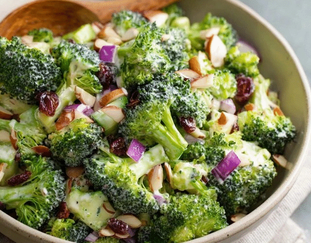 Broccoli salad with almonds in a large white serving bowl, bright green raw broccoli florets coated in creamy ivory dressing, crispy crumbled bacon bits, ruby red cranberries, golden toasted almond slivers, red onion specks