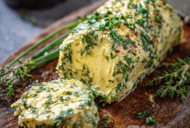Herb compound butter rolled into a log on parchment paper, soft pale-yellow butter studded with bright green chopped parsley, chives and thyme, garlic specks visible, lemon zest flecks, sliced into rounds on a wooden board