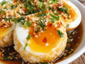 Soy sauce eggs with rice in a black bowl, halved jammy eggs with bright orange runny yolks over fluffy white rice, glossy soy-mirin marinade pooling, scallions and sesame seeds garnish, chili flakes on top