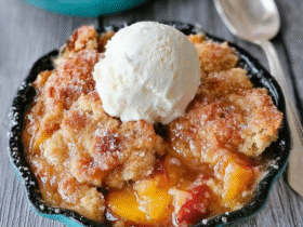 Peach cobbler in a baking dish with golden biscuit topping caramelized brown, juicy bubbling peach filling visible through cracks, large scoop of vanilla ice cream melting on top, cinnamon dust, fresh peach slices garnish