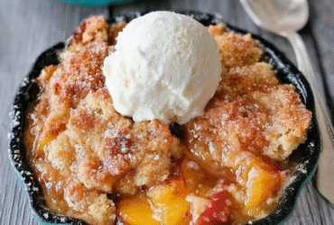 Peach cobbler in a baking dish with golden biscuit topping caramelized brown, juicy bubbling peach filling visible through cracks, large scoop of vanilla ice cream melting on top, cinnamon dust, fresh peach slices garnish