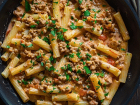 Creamy pasta with ground meat in a white bowl, penne pasta coated in pink tomato-cream sauce, browned ground beef visible throughout, melted parmesan strands, fresh basil leaves garnish, parmesan grated on top