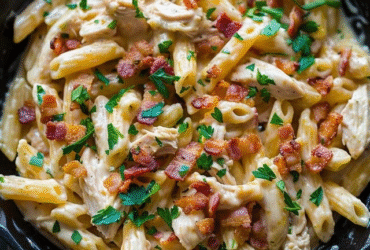Chicken bacon pasta in a white serving bowl, penne tubes coated in creamy garlic-parmesan sauce, golden grilled chicken strips, crispy crumbled bacon scattered throughout, fresh parsley, parmesan flakes on top