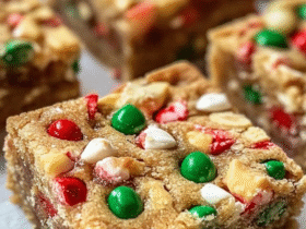 Holiday cookie bars cut into squares on parchment paper, soft buttery sugar cookie base with thick swirly cream cheese frosting layer on top, colorful red green and white festive sprinkles covering everything, cookie tin with more bars beside