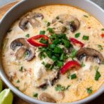 Slow cooker tom kha gai soup in a deep bowl, creamy coconut broth, tender chicken pieces, mushrooms, fresh cilantro and Thai chili, lime wedge on the rim, soft natural light