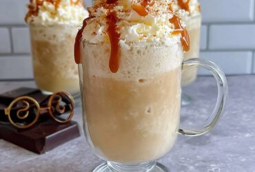 Two tall mugs of frozen butterbeer slush with thick foamy butterscotch whipped topping overflowing, dripping butterscotch drizzle down the sides, magical autumn light, Harry Potter themed