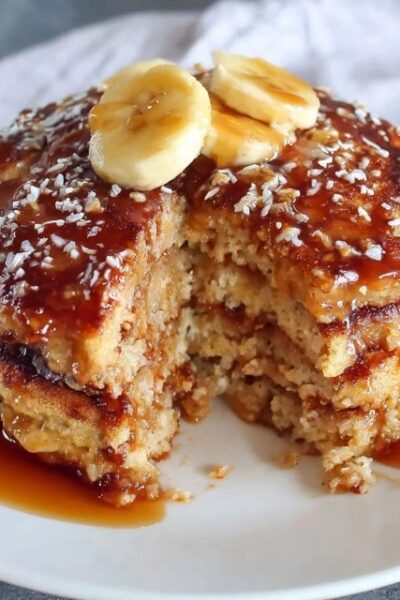 Stack of fluffy oatmeal pancakes on a wooden plate, golden brown edges, pat of butter melting on top, drizzle of maple syrup running down the sides, fresh blueberries scattered around, bright morning light