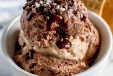 Two scoops of homemade chocolate ice cream in a glass coupe, glossy dark chocolate surface, slight melt drip showing creamy interior, chocolate sauce drizzle and chocolate shavings on top
