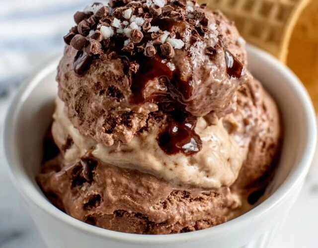 Two scoops of homemade chocolate ice cream in a glass coupe, glossy dark chocolate surface, slight melt drip showing creamy interior, chocolate sauce drizzle and chocolate shavings on top