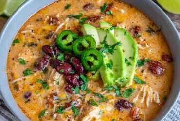 Creamy chicken enchilada soup in a black bowl, deep orange-red broth, shredded chicken, black beans, corn, melted cheese, topped with crispy tortilla strips, avocado, cilantro, lime wedge, sour cream dollop