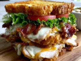 Crispy ultimate club sandwich cut diagonally and held together with frilly toothpicks, three layers of toasted bread, layers of crispy bacon, sliced turkey, lettuce, tomato, avocado, fanned out on a wooden board with potato chips and a pickle spear