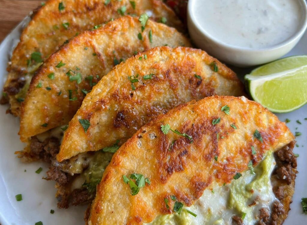 Crispy ground beef tacos lined up on a wooden board, golden crunchy taco shells filled with seasoned ground beef, shredded lettuce, diced tomato, cheddar cheese, dollop of sour cream, lime wedges and pickled jalapeños on the side