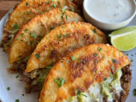 Crispy ground beef tacos lined up on a wooden board, golden crunchy taco shells filled with seasoned ground beef, shredded lettuce, diced tomato, cheddar cheese, dollop of sour cream, lime wedges and pickled jalapeños on the side