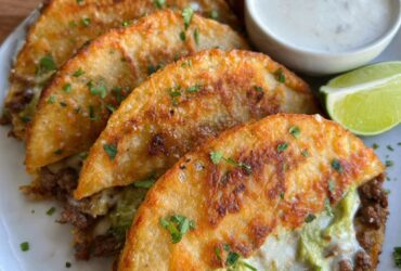 Crispy ground beef tacos lined up on a wooden board, golden crunchy taco shells filled with seasoned ground beef, shredded lettuce, diced tomato, cheddar cheese, dollop of sour cream, lime wedges and pickled jalapeños on the side