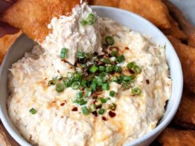 Creamy crab dip in a cast iron skillet, golden bubbly cheese top with browned spots, fresh chives and Old Bay seasoning sprinkled on top, served with crackers, baguette slices, and celery sticks around it