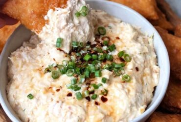 Creamy crab dip in a cast iron skillet, golden bubbly cheese top with browned spots, fresh chives and Old Bay seasoning sprinkled on top, served with crackers, baguette slices, and celery sticks around it