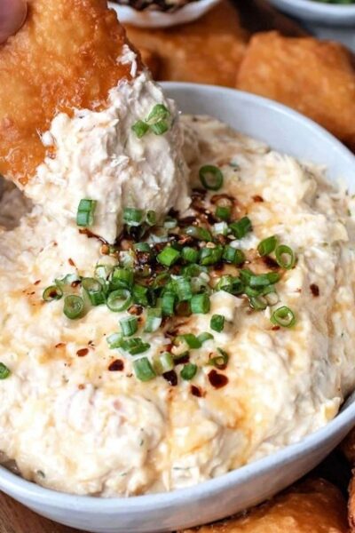 Creamy crab dip in a cast iron skillet, golden bubbly cheese top with browned spots, fresh chives and Old Bay seasoning sprinkled on top, served with crackers, baguette slices, and celery sticks around it