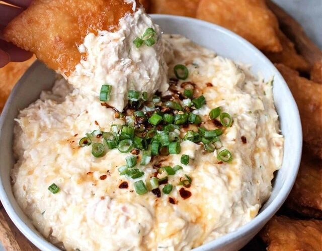 Creamy crab dip in a cast iron skillet, golden bubbly cheese top with browned spots, fresh chives and Old Bay seasoning sprinkled on top, served with crackers, baguette slices, and celery sticks around it