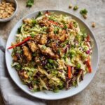 Asian chicken salad in a large white bowl, shredded purple and green cabbage, diced chicken, mandarin orange segments, sliced almonds, chow mein noodles, green onions, sesame ginger dressing drizzled on top