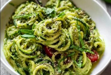 Zucchini noodles with pesto in a white bowl, fresh green spiralized zucchini coated in vibrant basil pesto, halved cherry tomatoes, pine nuts, shaved parmesan, fresh basil leaves on top
