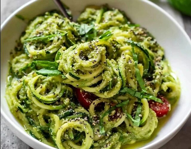 Zucchini noodles with pesto in a white bowl, fresh green spiralized zucchini coated in vibrant basil pesto, halved cherry tomatoes, pine nuts, shaved parmesan, fresh basil leaves on top