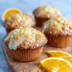 Orange muffins with glaze on a wire rack, tall domed muffin tops with white glaze drizzled over, fresh orange zest sprinkled on top, golden brown sides visible, halved muffin showing tender crumb