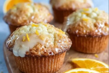 Orange muffins with glaze on a wire rack, tall domed muffin tops with white glaze drizzled over, fresh orange zest sprinkled on top, golden brown sides visible, halved muffin showing tender crumb