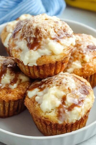 Banana cream cheese muffins on a wooden board, golden banana muffins with visible white cream cheese centers, sliced banana slices on top, sprinkle of brown sugar