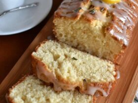 Lemon rosemary pound cake on a wooden serving board with a yellow honey-lemon glaze drizzled over the top, fresh rosemary sprigs garnishing, lemon zest curls, slice cut showing tight buttery crumb