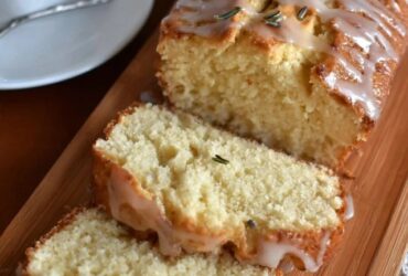 Lemon rosemary pound cake on a wooden serving board with a yellow honey-lemon glaze drizzled over the top, fresh rosemary sprigs garnishing, lemon zest curls, slice cut showing tight buttery crumb