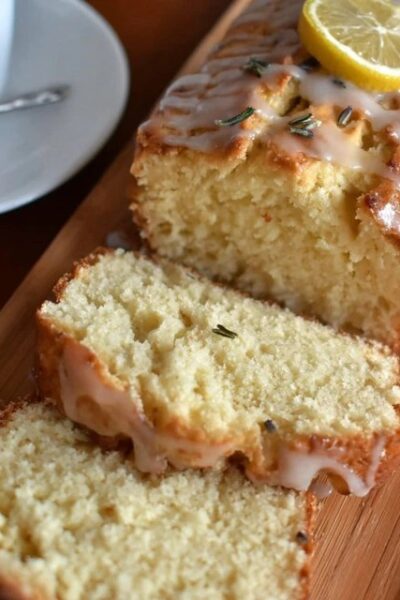 Lemon rosemary pound cake on a wooden serving board with a yellow honey-lemon glaze drizzled over the top, fresh rosemary sprigs garnishing, lemon zest curls, slice cut showing tight buttery crumb