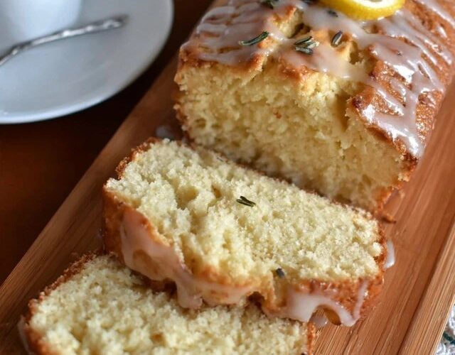 Lemon rosemary pound cake on a wooden serving board with a yellow honey-lemon glaze drizzled over the top, fresh rosemary sprigs garnishing, lemon zest curls, slice cut showing tight buttery crumb