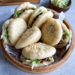 Bao buns with pork filling on a bamboo steamer, white pillowy soft buns split open with glossy hoisin-glazed pork belly slices, pickled cucumber, fresh cilantro, sliced scallions, sesame seeds