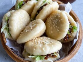 Bao buns with pork filling on a bamboo steamer, white pillowy soft buns split open with glossy hoisin-glazed pork belly slices, pickled cucumber, fresh cilantro, sliced scallions, sesame seeds