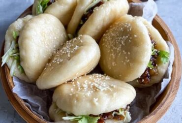 Bao buns with pork filling on a bamboo steamer, white pillowy soft buns split open with glossy hoisin-glazed pork belly slices, pickled cucumber, fresh cilantro, sliced scallions, sesame seeds