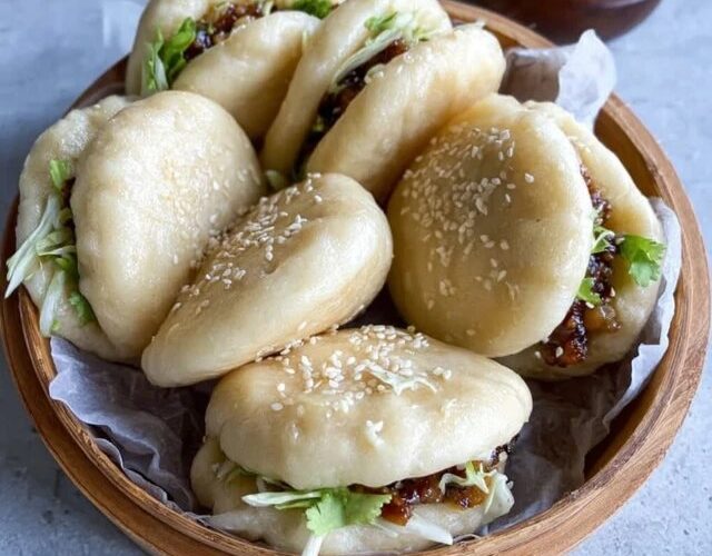 Bao buns with pork filling on a bamboo steamer, white pillowy soft buns split open with glossy hoisin-glazed pork belly slices, pickled cucumber, fresh cilantro, sliced scallions, sesame seeds