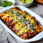 Cheese enchiladas in a casserole dish, rolled corn tortillas covered in deep red enchilada sauce and melted cheese, fresh cilantro, diced red onion on top, sour cream dollops, lime wedges, served in a Mexican ceramic dish